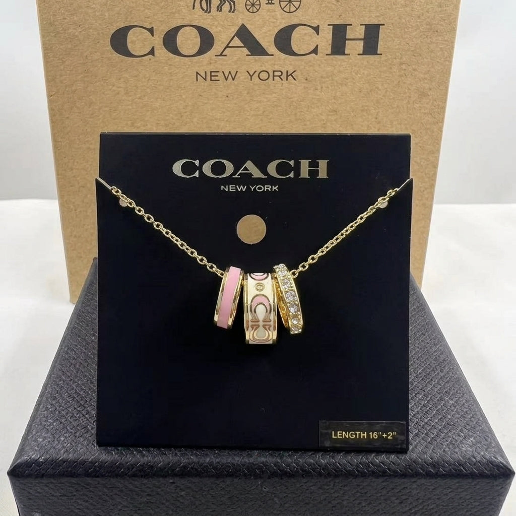 A Coach New York charm necklace displayed on a black branded card, with three dangling charms including a pink and gold patterned bead and a gold-toned chain, set against a kraft Coach box and dark presentation box.