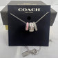 A Coach charm bracelet displayed on a black Coach New York card, with three silver-toned charms featuring the Coach logo and one pink accent charm, in front of the branded box.