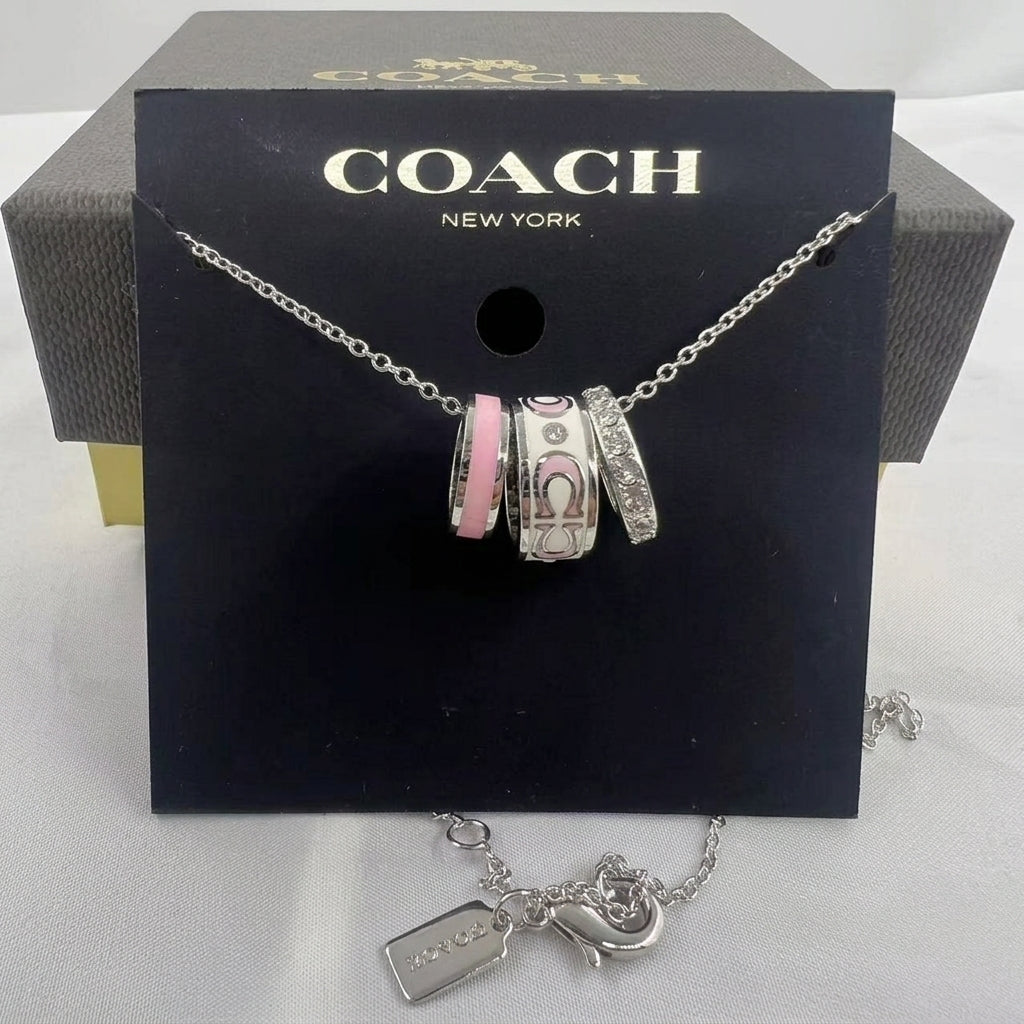 A Coach charm bracelet displayed on a black Coach New York card, with three silver-toned charms featuring the Coach logo and one pink accent charm, in front of the branded box.