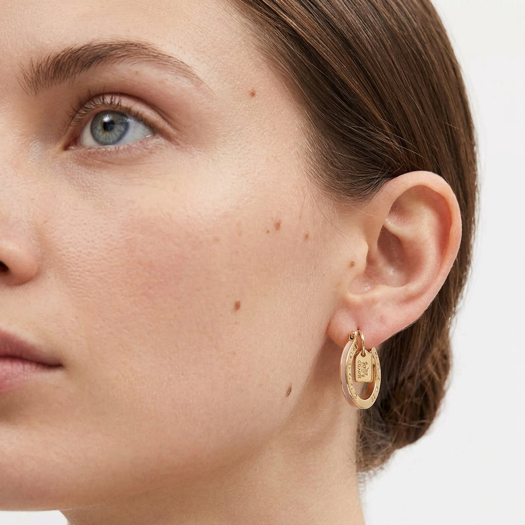 A close-up of a woman's ear wearing a gold hoop earring with a small rectangular charm, against a clean white background.
