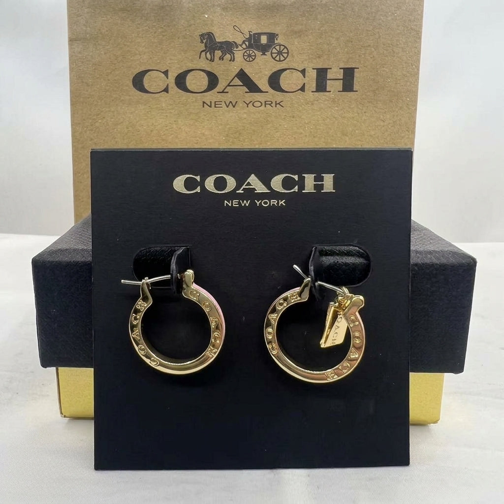A pair of gold-tone Coach hoop earrings with a small dangling tag, displayed on a black Coach New York card against a kraft Coach box and black gift box.