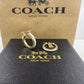 Gold and white Coach hoop earrings displayed on a black textured box with gold Coach branding, with a larger brown Coach box in the background.
