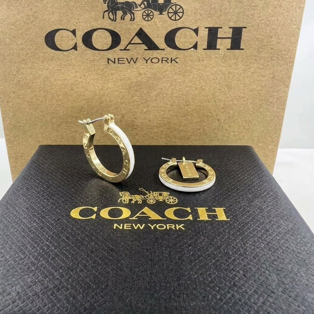 Gold and white Coach hoop earrings displayed on a black textured box with gold Coach branding, with a larger brown Coach box in the background.