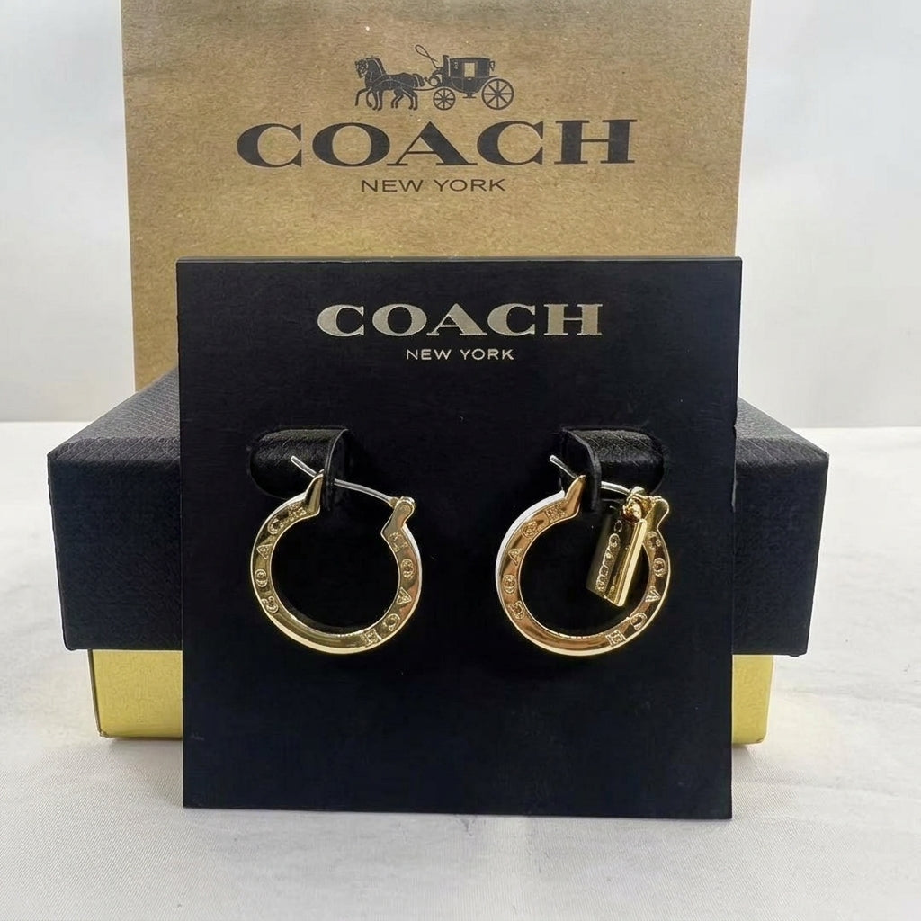 A pair of gold hoop earrings with Coach logo lettering, displayed on a black Coach New York card against a gold and black Coach box background.