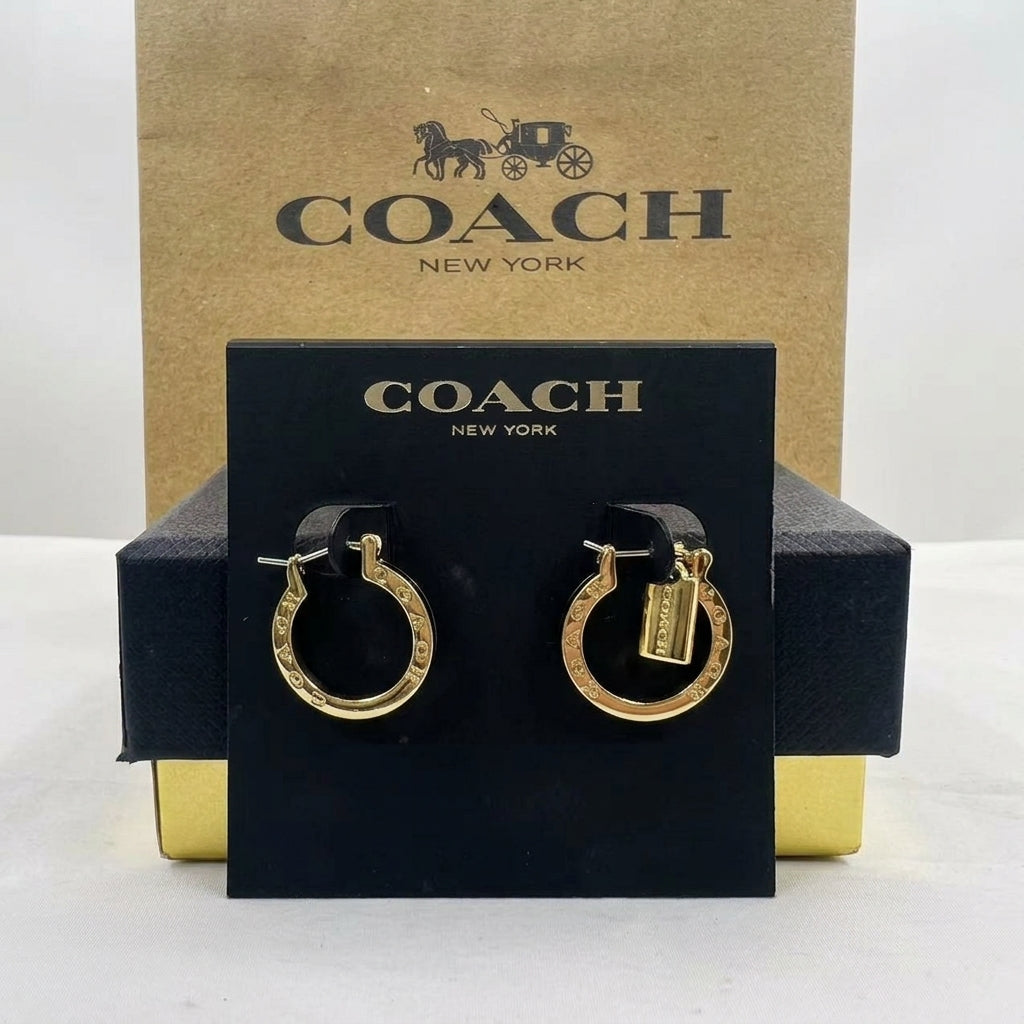 A pair of gold hoop earrings with Coach logo charms, displayed on a black branded card in front of a Coach New York box.