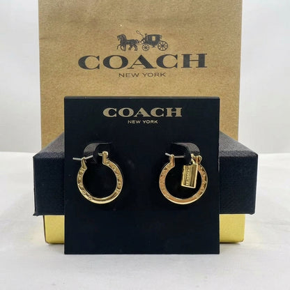 A pair of gold hoop earrings with Coach logo charms, displayed on a black branded card in front of a Coach New York box.