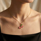 A close-up of a model's neck and upper chest wearing a delicate gold chain necklace with a cherry pendant. The pendant features pink enamel-filled cherries with green leaves, set against a warm, softly lit background.