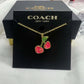 A Coach New York necklace with a gold-tone chain and a cherry pendant featuring pink enamel and green leaves, presented on a black Coach New York display card.