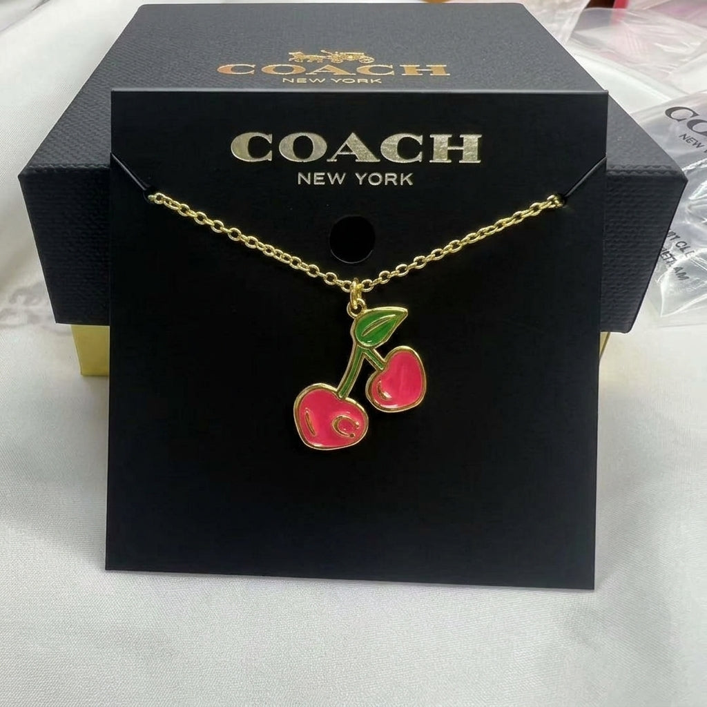 A Coach New York necklace with a gold-tone chain and a cherry pendant featuring pink enamel and green leaves, presented on a black Coach New York display card.
