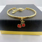A gold-plated bangle bracelet with a dangling cherry charm, displayed on a black textured jewelry box.