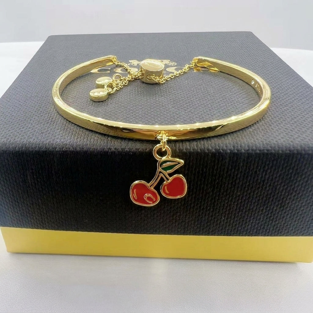 A gold-plated bangle bracelet with a dangling cherry charm, displayed on a black textured jewelry box.