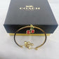 A gold-tone bangle bracelet with a cherry charm, displayed in front of its black Coach New York gift box on a white surface.