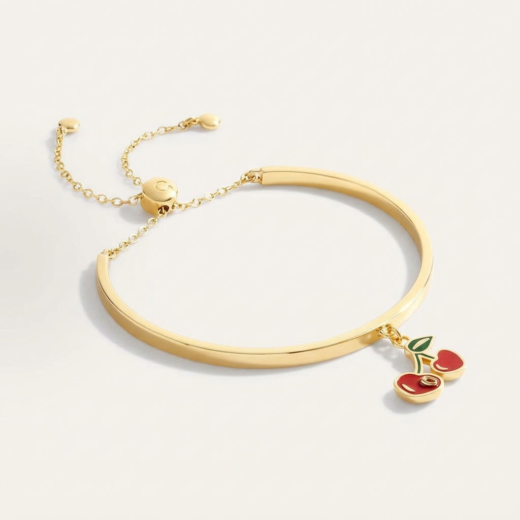 A gold-plated bangle bracelet with a delicate chain and slider clasp, featuring a dangling cherry charm with red enamel and a green leaf, set against a clean off-white background.