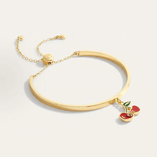 A gold-plated bangle bracelet with a delicate chain and slider clasp, featuring a dangling cherry charm with red enamel and a green leaf, set against a clean off-white background.