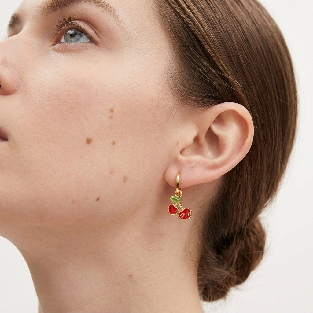 A close-up of a woman's ear wearing a small gold hoop earring with two red cherry charms and green leaves.