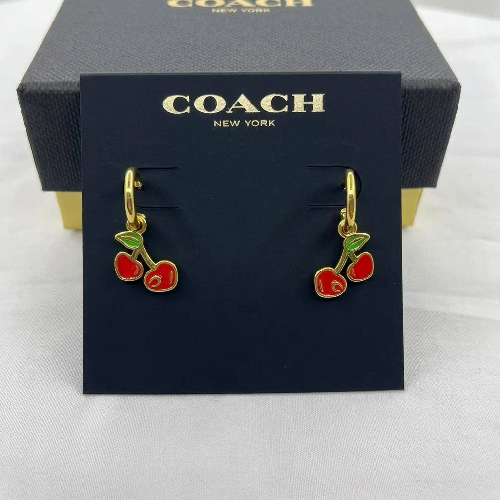 A pair of Coach New York cherry drop earrings with gold-tone metal and red and green enamel, displayed on a branded navy blue card with the Coach box behind it.