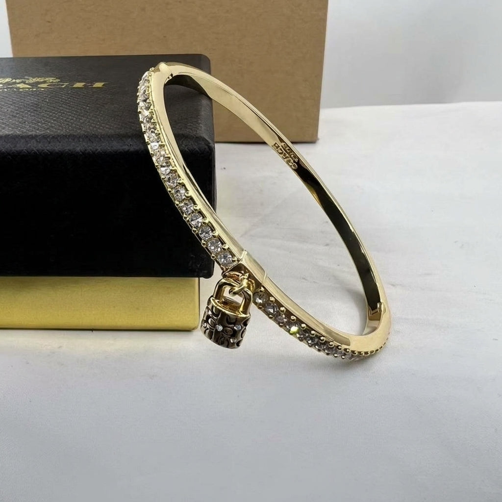 A gold-plated bangle bracelet with a row of clear pavé stones on one side and a small padlock charm, displayed next to a black jewelry box.