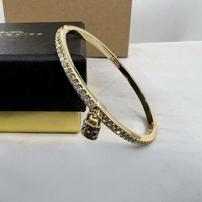 A gold-plated bangle bracelet with a row of clear pavé stones on one side and a small padlock charm, displayed next to a black jewelry box.