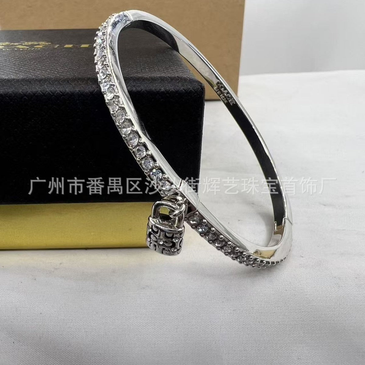 A silver-toned bangle bracelet with a row of clear stones and a small padlock charm, displayed on a white surface with a black jewelry box in the background.