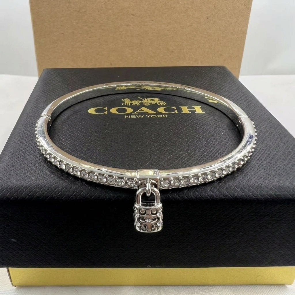 A silver-tone Coach bangle bracelet with a padlock charm, displayed on its black textured brand box.