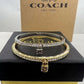 A pair of Coach bangle bracelets, one gold and one silver, both adorned with clear crystals and featuring a small padlock charm, displayed on a black box with a Coach New York box in the background.