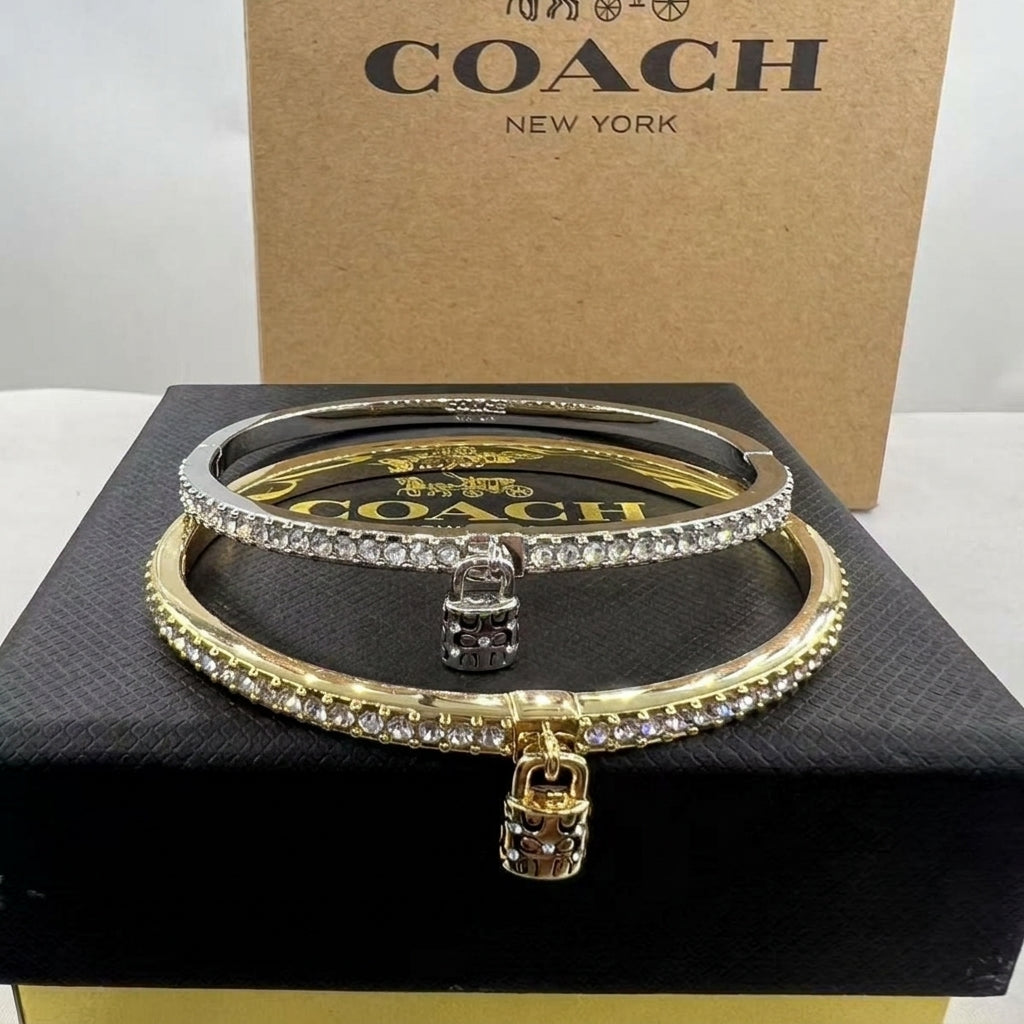 A pair of Coach bangle bracelets, one gold and one silver, both adorned with clear crystals and featuring a small padlock charm, displayed on a black box with a Coach New York box in the background.