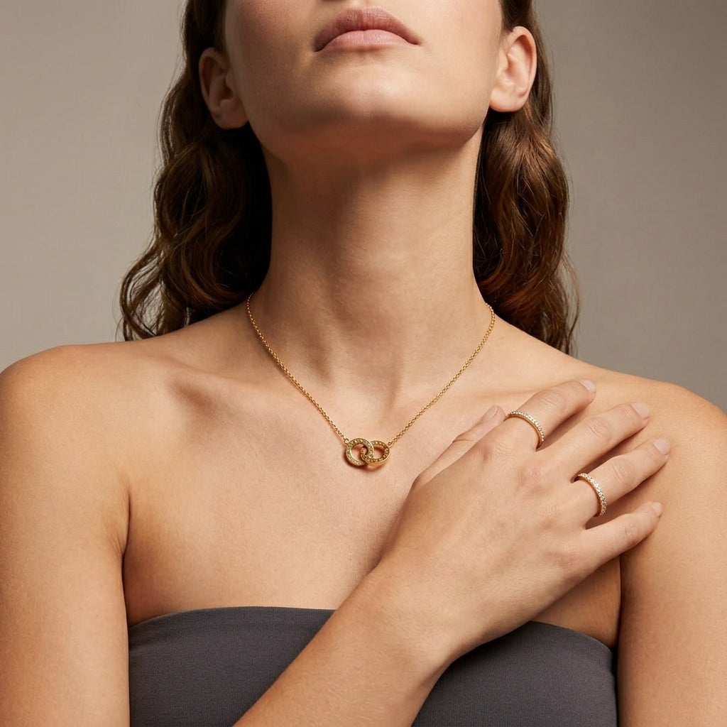 A close-up of a woman wearing a delicate gold necklace with a double-ring pendant and matching gold rings on her fingers, set against a neutral warm gray background.