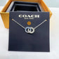 A Coach New York necklace displayed on a black branded card, with a gold-tone box behind it. The necklace has two interlocking circular charms with a blue and silver pattern, suspended from a silver-toned chain.