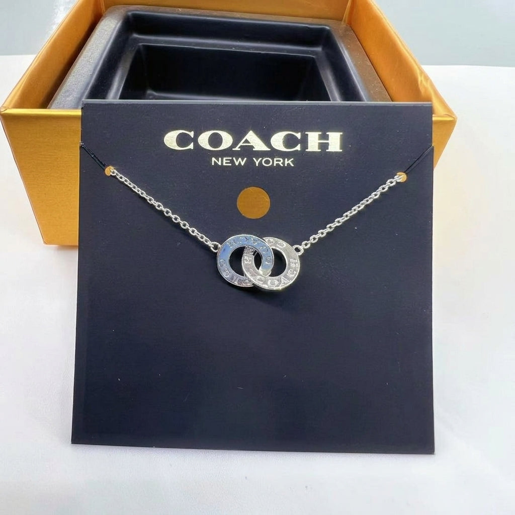 A Coach New York necklace displayed on a black branded card, with a gold-tone box behind it. The necklace has two interlocking circular charms with a blue and silver pattern, suspended from a silver-toned chain.