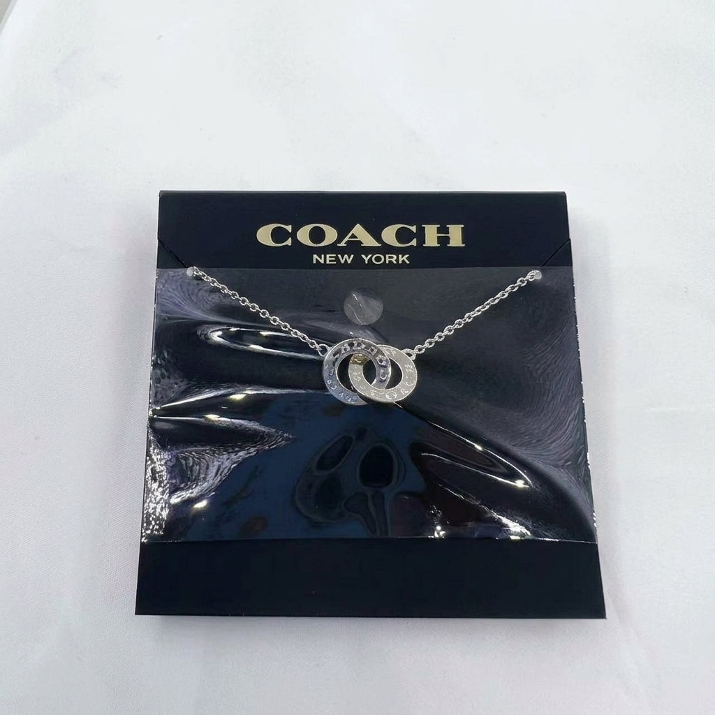 A silver-tone Coach New York interlocking logo pendant necklace on a chain, presented on its black branded display card.