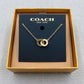 A gold-tone double-ring pendant necklace by Coach, displayed on its black branded card inside an open gold box on a gray textured background.
