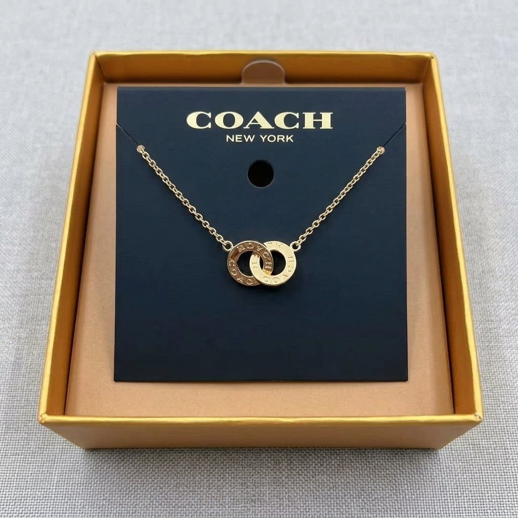 A gold-tone double-ring pendant necklace by Coach, displayed on its black branded card inside an open gold box on a gray textured background.