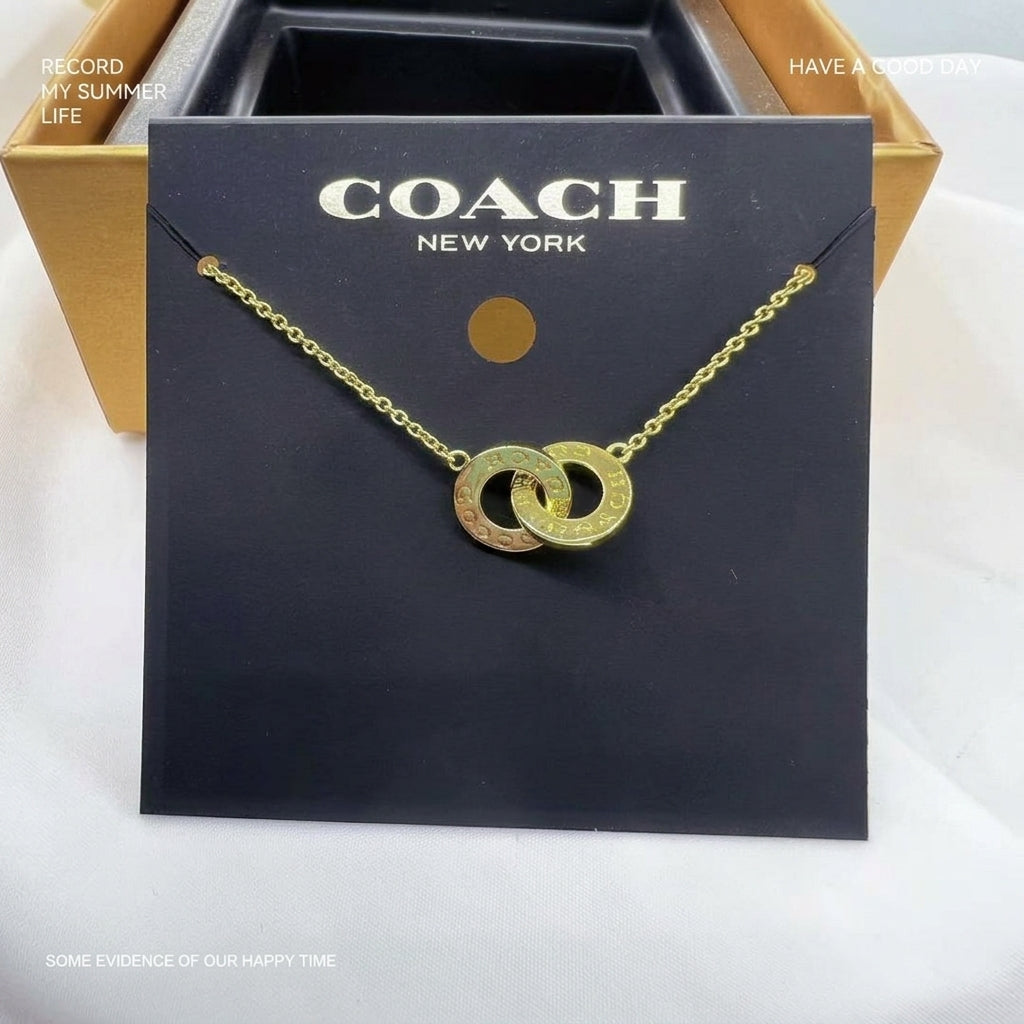 A gold-tone double interlocking ring necklace on a black Coach New York display card, with a gold and black Coach box partially visible behind it.