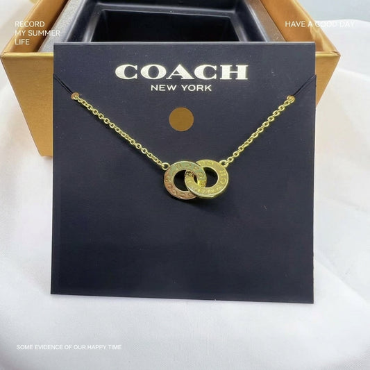A gold-tone double interlocking ring necklace on a black Coach New York display card, with a gold and black Coach box partially visible behind it.