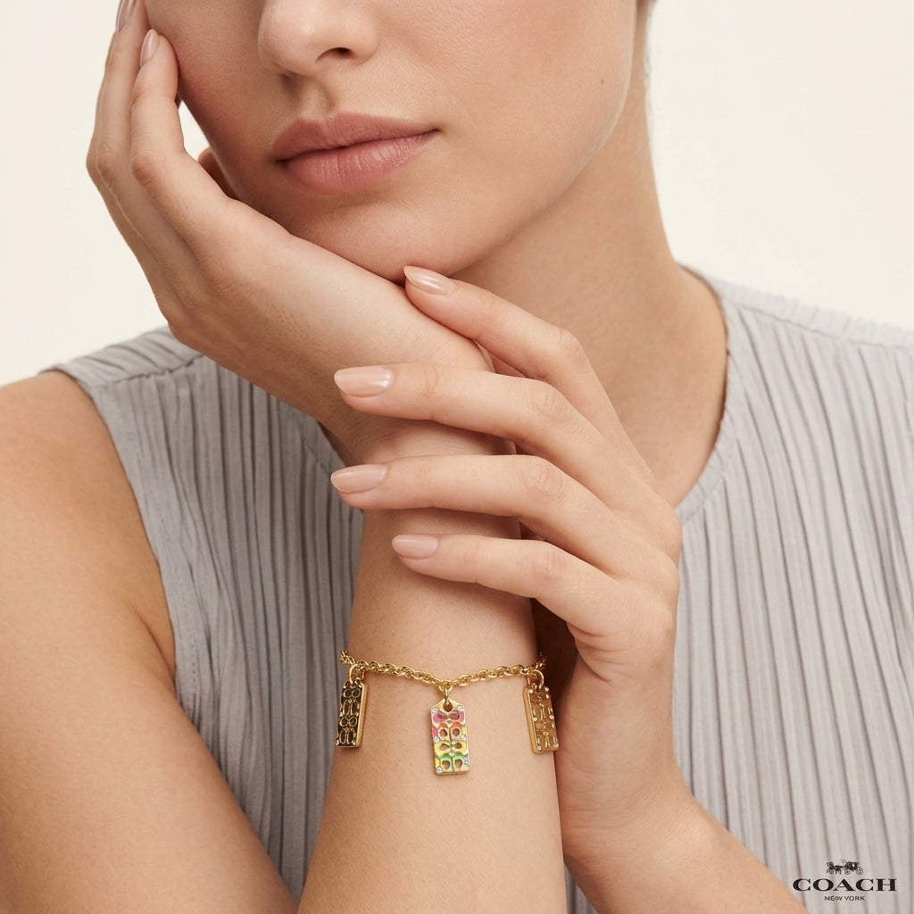 A close-up lifestyle shot of a model wearing a gold-tone charm bracelet with colorful rectangular charms, styled with a gray pleated top against a light beige background. The brand name 'COACH' is visible in the bottom right corner.