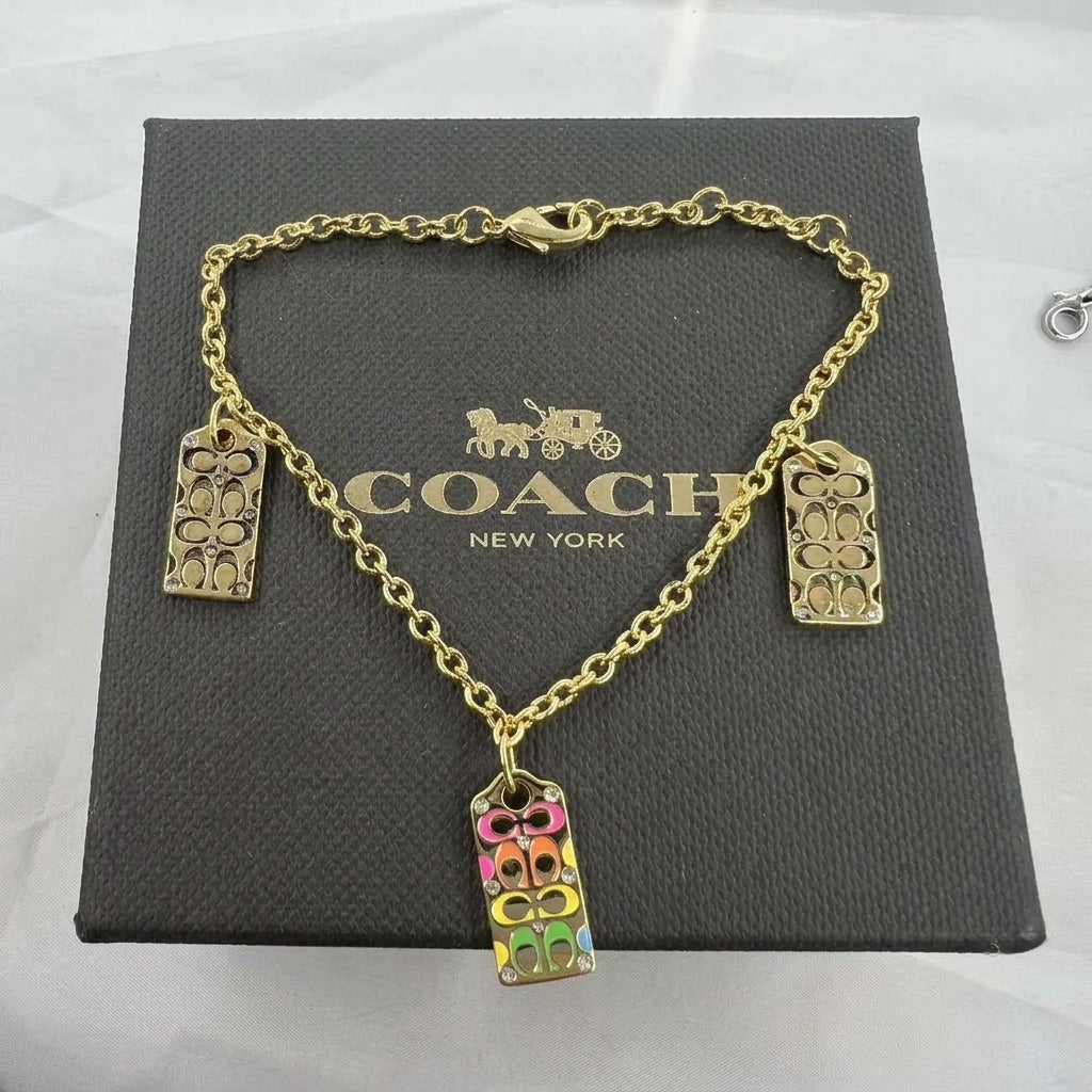 Gold-tone Coach charm bracelet displayed on a dark gray Coach New York gift box. The bracelet features a gold chain with three rectangular charms: two plain gold-tone floral signature charms and one colorful charm filled with pink, yellow, green, and gold-tone Coach logo details.