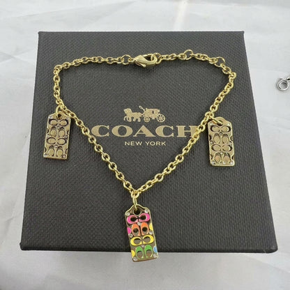 Gold-tone Coach charm bracelet displayed on a dark gray Coach New York gift box. The bracelet features a gold chain with three rectangular charms: two plain gold-tone floral signature charms and one colorful charm filled with pink, yellow, green, and gold-tone Coach logo details.