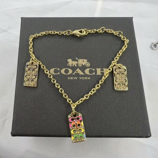 Gold-tone Coach charm bracelet displayed on a dark gray Coach New York gift box. The bracelet features a gold chain with three rectangular charms: two plain gold-tone floral signature charms and one colorful charm filled with pink, yellow, green, and gold-tone Coach logo details.
