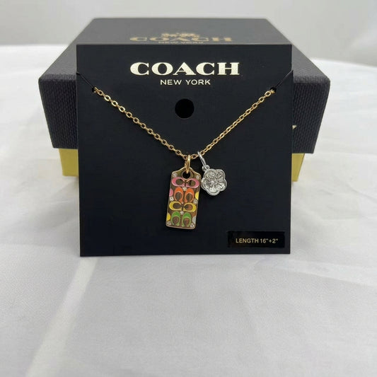 A Coach New York necklace displayed on a black branded card, with the necklace hanging in front of the box. The pendant is a colorful rectangular Coach logo charm with a silver floral charm, on a gold chain.