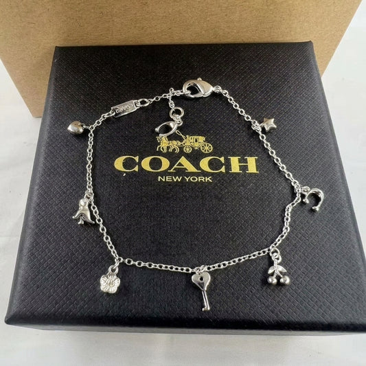 A silver-tone charm bracelet displayed on a black Coach New York box. The bracelet features multiple charms including a horse, a key, cherries, and a flower, attached to a delicate chain with a lobster clasp.