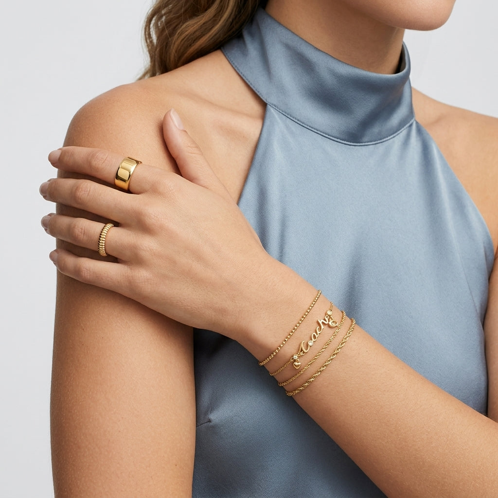 A close-up of a model's shoulder and arm wearing multiple gold jewelry pieces, including a wide band ring, a textured ring, and three delicate name bracelets with the name 'Emily'. The model is wearing a high-neck dusty blue satin top against a clean white background.