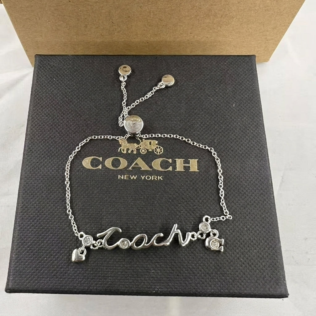 A silver-tone Coach nameplate bracelet with a carriage charm and small rhinestone accents, displayed on a black Coach New York presentation box.