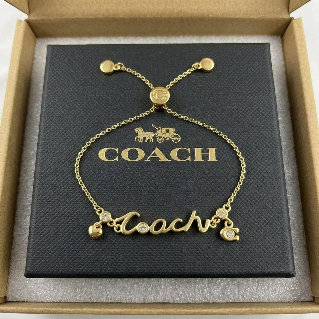 A gold-tone Coach nameplate bracelet with crystal accents, presented on its black branded box inside an open brown presentation box.