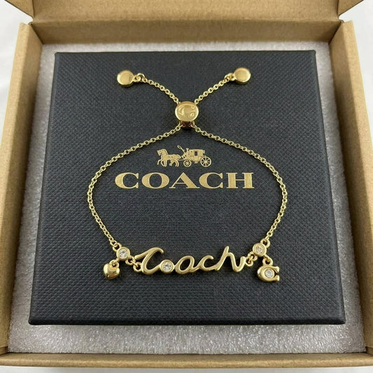 A gold-tone Coach nameplate bracelet with crystal accents, presented on its black branded box inside an open brown presentation box.