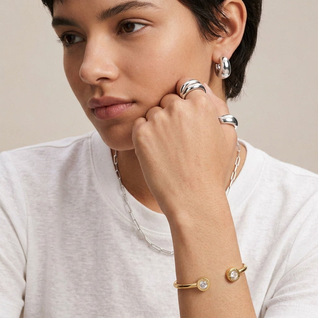 A close-up lifestyle shot of a model wearing a collection of minimalist jewelry, including silver rings, a silver hoop earring, a silver chain necklace, and a gold bangle bracelet with clear crystal accents, against a soft beige background.