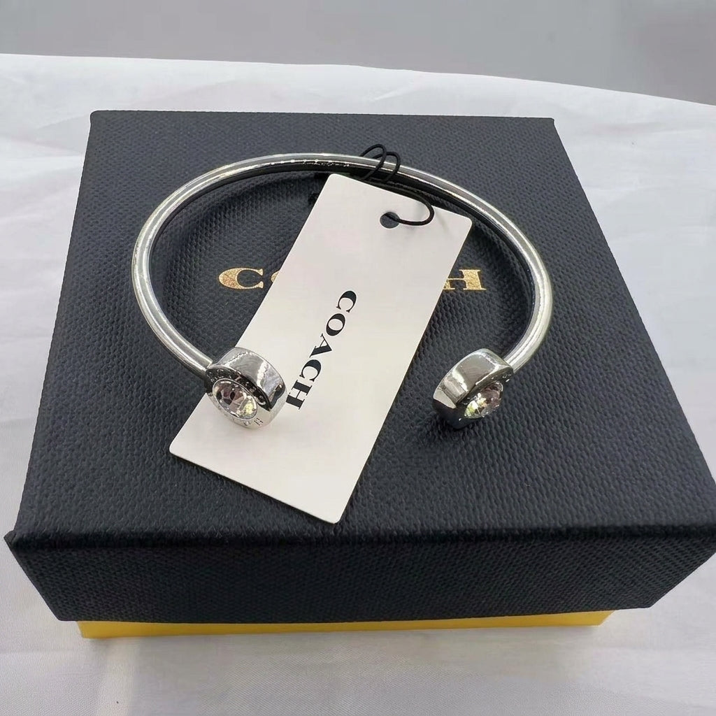 A silver-tone cuff bracelet with two crystal-encrusted end caps, displayed on top of a black Coach box with a white Coach hangtag.