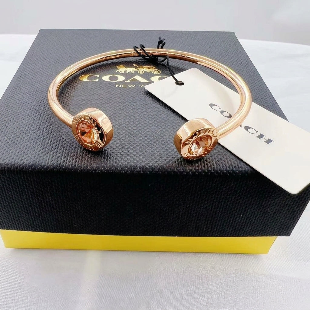 A Coach cuff bracelet in rose gold with two round crystal-encrusted end caps, displayed on top of a black Coach box with a white brand tag.