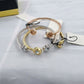 Three slim bangle bracelets in rose gold, yellow gold, and silver, each featuring a small faceted crystal bead at the end, arranged on a light gray fabric background with paper tags.
