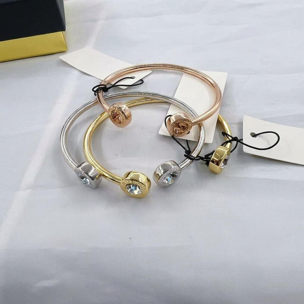 Three slim bangle bracelets in rose gold, yellow gold, and silver, each featuring a small faceted crystal bead at the end, arranged on a light gray fabric background with paper tags.