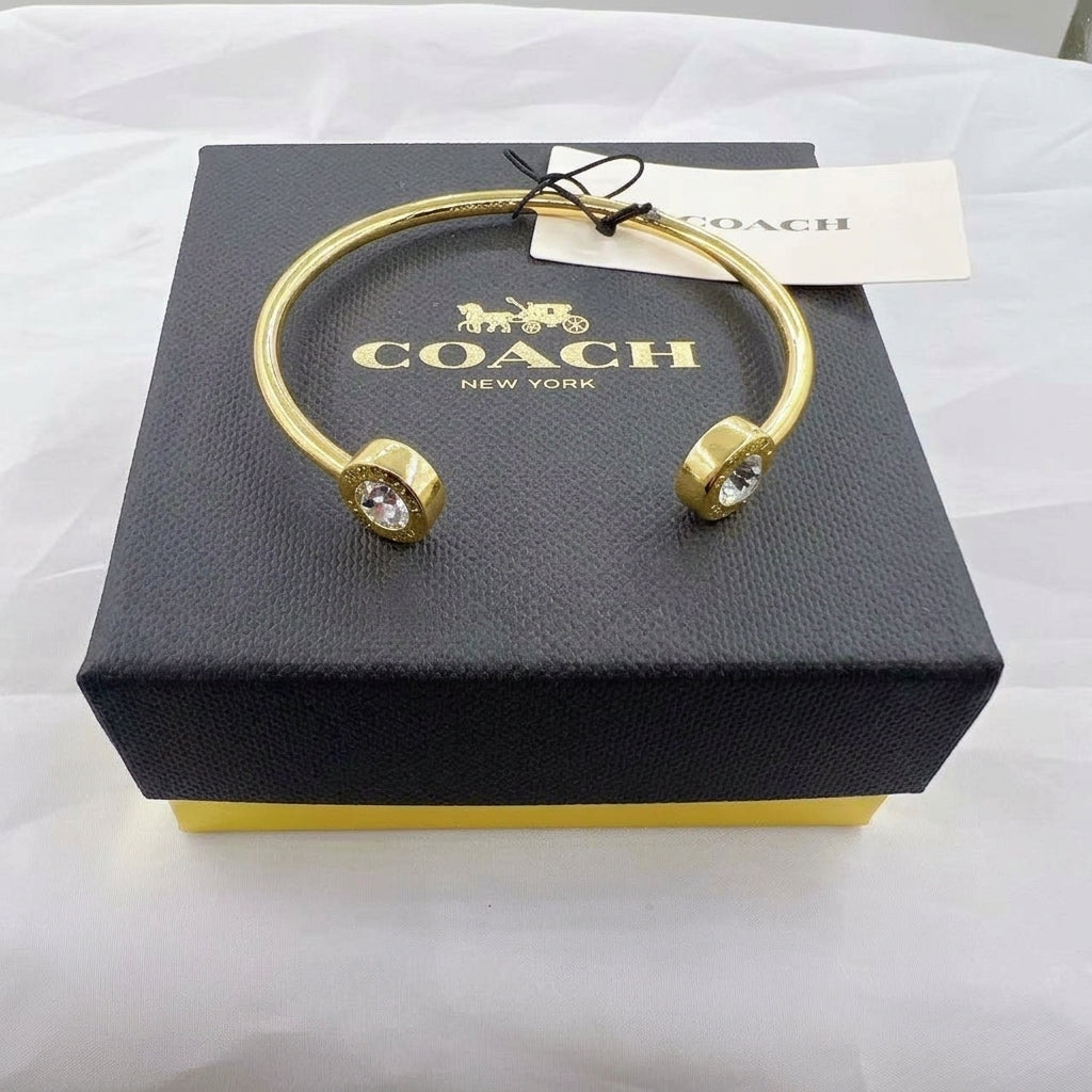 A gold-tone cuff bangle bracelet with clear crystal accents on both ends, displayed on top of a black Coach New York gift box with a white hangtag.